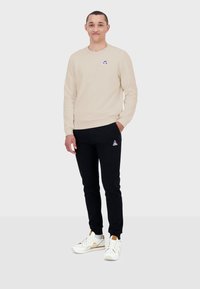 Sudadera beige con cuello redondo, pantalones de chándal negros y zapatillas blancas con acentos dorados. Presenta un pequeño logo bordado en cada uno.