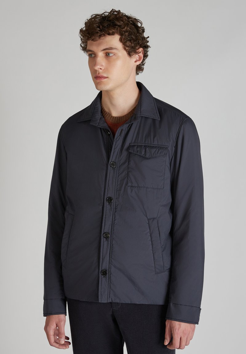 Granatowa, lekka kurtka z zapięciem na guziki, kieszenią na piersi i płaskim kołnierzykiem, wykonana z gładkiego, fakturowanego materiału.