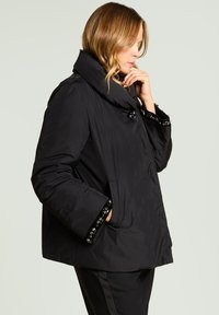 Abrigo acolchado negro con cuello ancho, con detalles decorativos en los puños y cierre frontal con botones. Incluye dos bolsillos laterales.