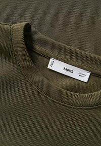 Tissu en coton vert olive texturé avec un col rond et des coutures visibles. L'étiquette attachée indique la marque "MNG" et les détails de la taille.