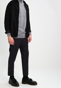 Giacca nera con colletto sopra un maglione di maglia grigia, abbinata a pantaloni scuri con fantasia e scarpe nere con suola spessa.