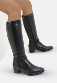Botas de cuero negras hasta la rodilla con puntera afilada, tacón bajo en bloque, cremallera lateral y una correa en el tobillo con tachuelas decorativas. Textura suave.