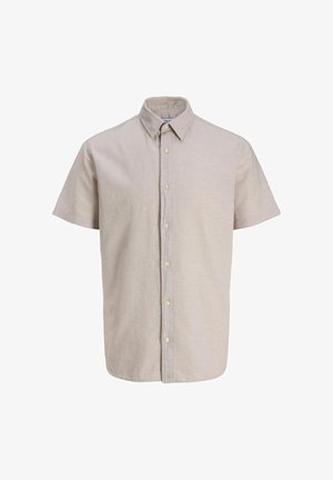 Adolescente con capelli ricci in piedi e sorridente, indossa una camicia beige a maniche corte con bottoni sopra una maglietta bianca, pantaloni neri e scarpe da ginnastica bianche.