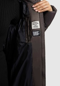 Binnenkant van de pufferjack, donkergrijze stof met een gequilt ontwerp. Labels vermelden "Een warme omhelzing gemaakt van stof" en "Khujo origineel met veranderende draden."