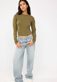 Olīvu zaļš garām piedurknēm īsais tops ar pieguļošu dizainu, apvienots ar gaiši ziliem platām denim biksēm. Redzama izteikta šuvju detaļa.