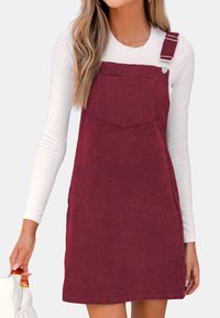Bordeaux corduroy jurk met verstelbare bandjes, voorzak en rechte snit. Gecombineerd met een witte blouse met lange mouwen.