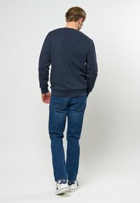 Marineblauwe sweatshirt met een ronde hals, geribbelde mouwen en onderrand, gecombineerd met blauwe spijkerbroek en witte sneakers, gezien van achteren.