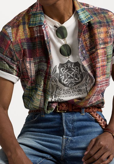 Daudzkrāsains, rakstains īsām Piedurknēm krekls, kas uzlikts virs balta T-krekla ar grafiku, apvienots ar zilu džinsu biksēm un brūnu pinumu jostu.