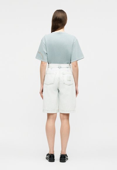 Γυναίκα με ανοιχτό μπλε πουκάμισο με κοντά μανίκια, σορτς βερμούδα τζιν ανοιχτού χρώματος με ψηλή μέση και μαύρα παπούτσια, στέκεται γυρισμένη προς τα πίσω σε λευκό φόντο.