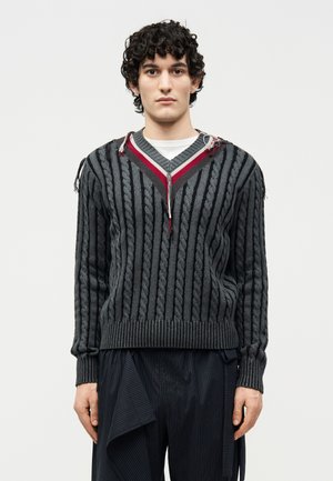 Νεαρός με σγουρά σκούρα μαλλιά που φοράει γκρι πλεκτό πουλόβερ V-λαιμόκοψη με κόκκινες ρίγες και σκούρο φαρδύ παντελόνι με λεπτές ρίγες.