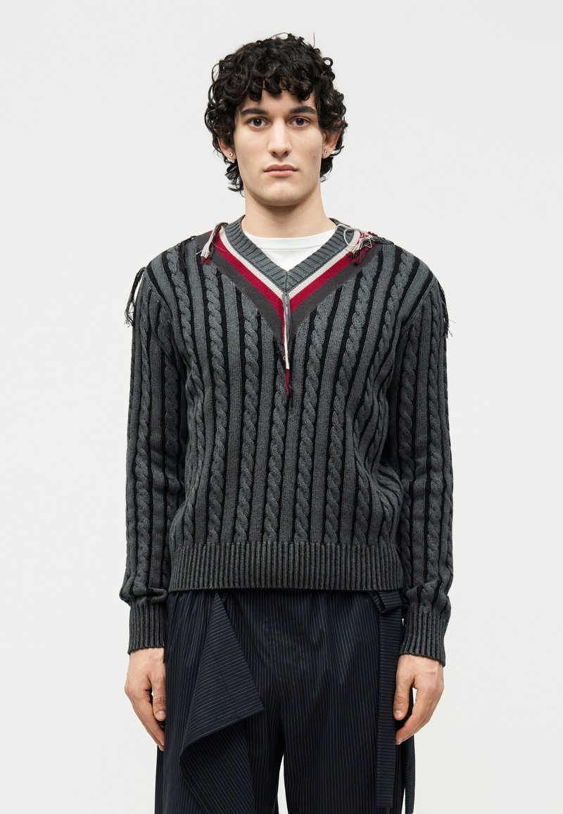 Νεαρός με σγουρά σκούρα μαλλιά που φοράει γκρι πλεκτό πουλόβερ V-λαιμόκοψη με κόκκινες ρίγες και σκούρο φαρδύ παντελόνι με λεπτές ρίγες.