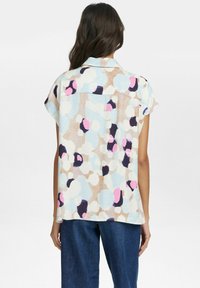 Blouse à manches courtes avec un motif multicolore de formes bleues, roses et noires sur un fond beige ; tissu lisse avec une coupe décontractée.