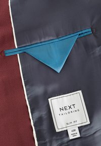 Sötét tengerészkék, bordó mintás, testre szabott zakóbélés. Égkék zseb és egy "NEXT TAILORING SLIM FIT" feliratú címke található rajta.