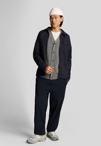 Giacca softshell navy sopra un cardigan grigio, t-shirt bianca, pantaloni scuri e sneakers grigie. Completa il look un berretto bianco a maglia a coste.