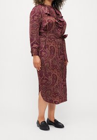 Vestito lungo color bordeaux con un motivo paisley, dettagli a volant sul collo, cintura con lacci in vita e maniche lunghe. Abbinato a mocassini neri.