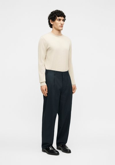 Άνδρας με σγουρά μαλλιά που φοράει μπεζ πουκάμισο με μακριά μανίκια, μαύρο παντελόνι με φαρδιά πόδια και μαύρα επίσημα παπούτσια, στέκεται σε λευκό φόντο.