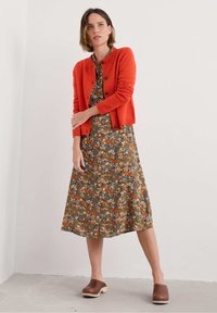 Cardigan en tricot orange sur une robe à motif floral marron. Sabots marron à semelles claires. Texture lisse, détails de boutons sur le cardigan.