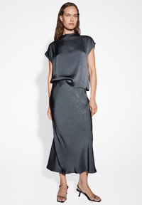 Zwarte satijnen twee-delige outfit met een losse top met korte mouwen en een vloeiende, enkel-lange rok met subtiele textuur. Eenvoudige zwarte sandalen.