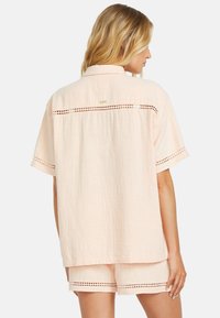 Kurzärmliges, hellpfirsichfarbenes Shirt mit lockerer Passform, strukturiertem Stoff und Spitzenbesatzdetails an Rücken und Ärmeln. Kombiniert mit passenden Shorts.