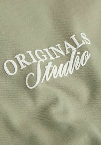 Φωτεινό πράσινο ύφασμα με λευκή κεντημένη επιγραφή "ORIGINALS Studio" σε ρευστή γραφή. Λείου υφής με υποβαθμισμένο σχέδιο.