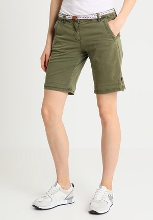 Shorts en coton vert olive avec une coupe ajustée, dotés de poches latérales et d'une taille décorative. Associés à des chaussures de sport blanches et grises.