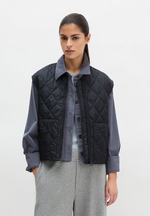 Donna che indossa un gilet trapuntato nero sopra una camicia grigia con bottoni e pantaloni grigio chiaro, in piedi davanti a uno sfondo bianco semplice.