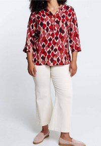 Donna che indossa una blusa a righe rossa e rosa, pantaloni beige a gamba larga e scarpe slip-on rosa chiaro, in piedi contro uno sfondo neutro.