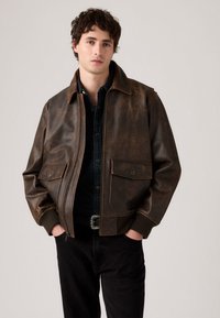 Brown leather jacket with front zip closure, two buttoned flap pockets, and ribbed cuffs. Worn over a black shirt and dark jeans.