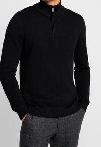 Maglione nero con zip e collo alto, con un motivo a maglia testurizzata, polsini e orlo a coste. Abbinato a pantaloni grigi a fantasia.