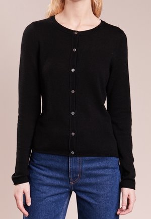 Frau trägt eine taillierte schwarze, geknöpfte Strickjacke mit langen Ärmeln und blaue Jeans, steht vor einem schlichten beigen Hintergrund.