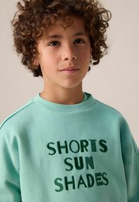 Sweat-shirt bleu clair à manches courtes, avec un texte vert foncé "SHORTS SUN SHADES". Tissu doux avec une texture subtile.