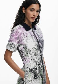 Robe à manches courtes avec un zip à l'avant, un col noir et un imprimé graphique comportant des textures violettes, vertes et noires sur fond blanc.