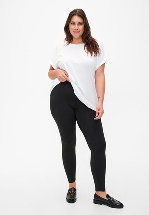 Svarta leggings med en slät textur i kombination med en oversized vit t-shirt. Modellen har även på sig svarta loafers med gyllene detaljer.