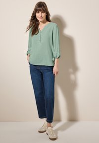 Maglietta verde chiaro a maniche lunghe, con bottoni e scollatura a V, abbinata a jeans boyfriend blu scuro e sneakers color crema.