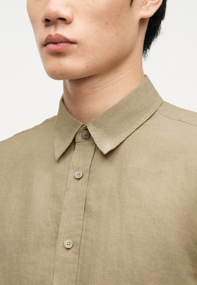 Νεαρό άτομο που φοράει μια μπεζ λινή πουκαμίσα με κουμπιά και μυτερό γιακά, εμφανίζεται από τη μύτη έως το πάνω μέρος του στήθους, με απλό φόντο.