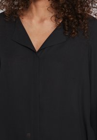 Blouse noire avec un éclat subtil, présentant un décolleté en V, une coupe ample et des détails de couture minimaux. Tissu doux avec une texture lisse.