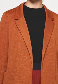 Personne portant un blazer texturé ouvert couleur orange brûlé sur un haut noir et une jupe rouge foncé sur fond blanc.