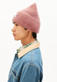 Roze gebreide beanie met een puntige bovenkant, met een textuurpatroon. Draagt een spijkerjack en een groen-wit geruit shirt.