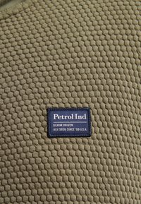 Strukturerad olivgrön tyg med ett 3D-bihoneycomb-mönster. En rektangulär blå etikett ger texten "Petrol Ind, Denim Driven, REF.5026 SINCE '89 U.S.A."