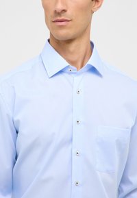 Camicia azzurra chiara con colletto a punta, tasca frontale e cinque bottoni lucidi, realizzata in un tessuto morbido.