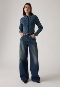 Chemise en denim avec col et boutons sur le devant associée à un pantalon en denim à jambes larges, tous deux en bleu foncé avec dégradé. Ceinture et bottes noires.