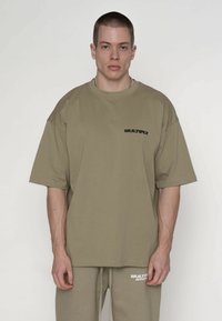 Oversized kortærmet T-shirt i olivengrøn bomuld med en sort "MULTIPLY"-logo på brystet. Har en afslappet pasform og rund halsudskæring.