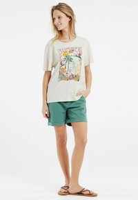 Camiseta gráfica de color crema con un estampado de temática veraniega, combinada con pantalones cortos en tono teal y chanclas marrones, con fondo blanco.