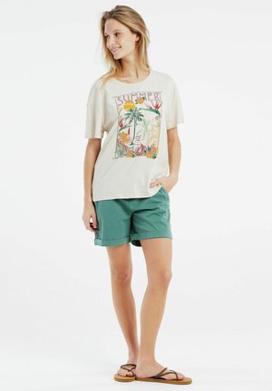T-shirt graphique crème avec un motif d'été, associé à un short teal et des tongs marron, se tenant devant un fond blanc.