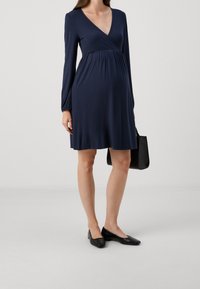 Modrá těhotenská šaty po kolena s výstřihem do V a dlouhými rukávy, vyrobené z hladkého materiálu. Doplněné černou texturovanou kabelkou a balerínami.