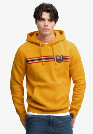 Jeune homme portant un sweat à capuche jaune moutarde avec des rayures rouges, noires et beiges et un logo circulaire de montagne sur la poitrine, associé à un jean bleu.