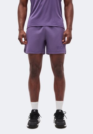 Homme portant un short et un haut de sport violets, des chaussettes blanches montantes et des chaussures de course Adidas noires, debout devant un fond clair uni.