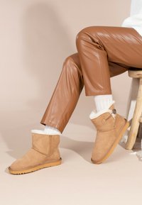Botas de tobillo de ante en color marrón claro con un forro de borrego blanco, un pequeño botón decorativo en el lateral y una suela de goma texturada.