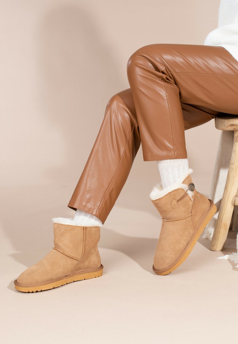 Botas de tobillo de ante en color marrón claro con un forro de borrego blanco, un pequeño botón decorativo en el lateral y una suela de goma texturada.