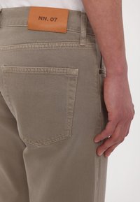 Pantalon beige avec poche arrière et patch en cuir marron marqué "NN. 07", porté par une personne ayant la main posée à côté de la jambe.